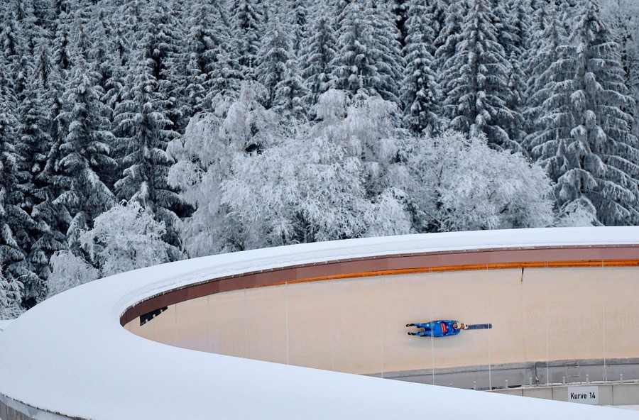 A pair of lugers make a run on a steep, curved track in a snowy forest.