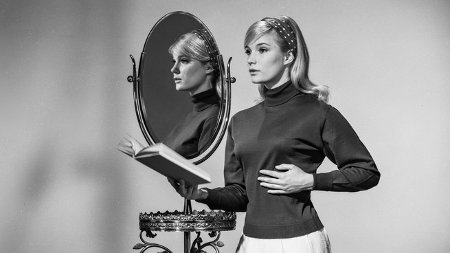 A vintage black-and-white photo of a woman holding a book in front of a mirror