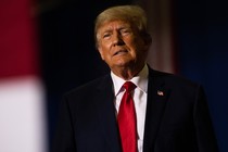 Color photograph of Donald Trump talking, wearing a black suit with a red tie.