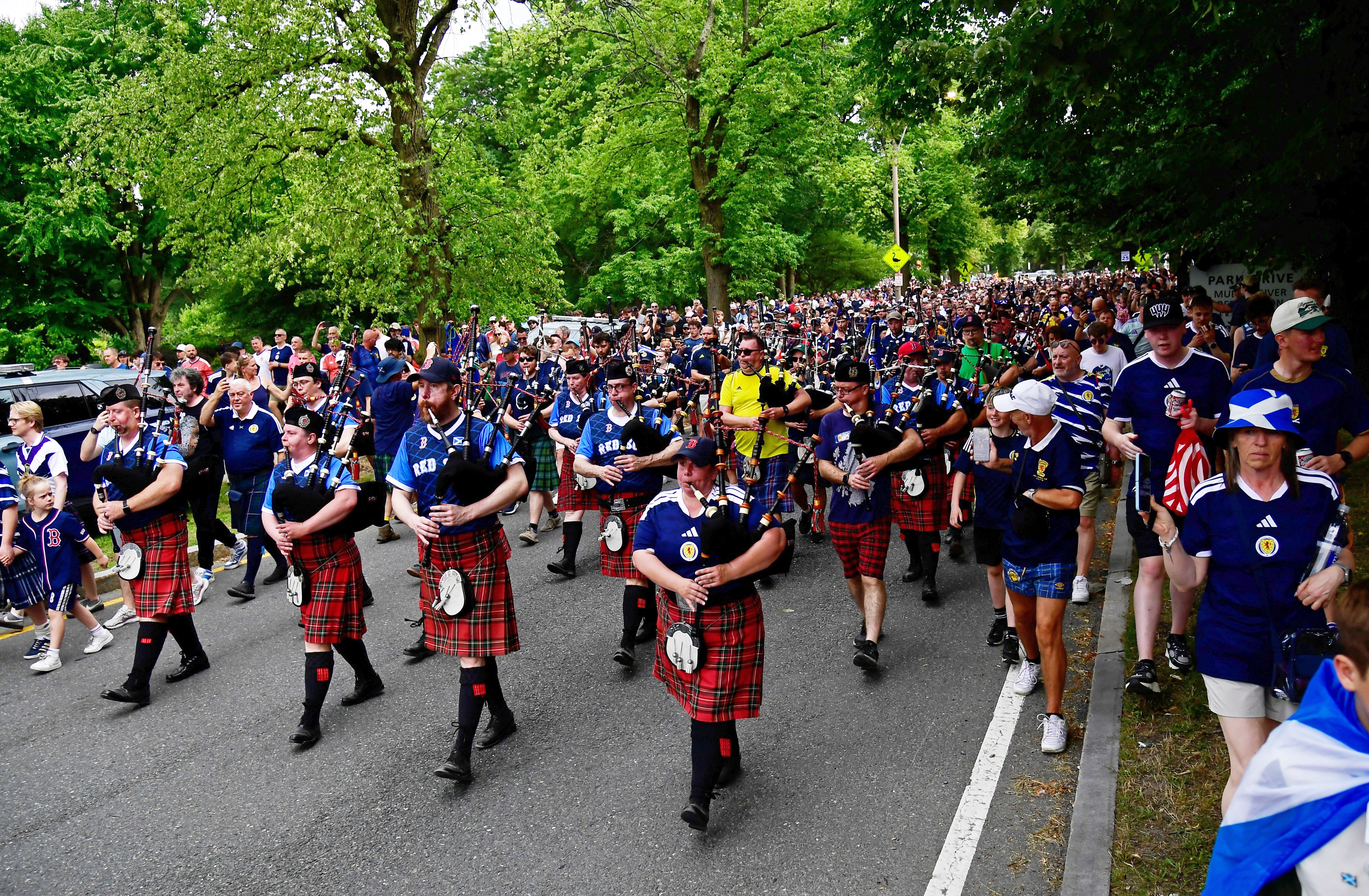 Scotland fans wearing kilts and playing the bagpipes march down a tree-line street toward Boston's Fenway Park.