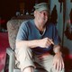 A white man sits in a chair holding a cigarette.
