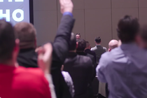 A crowd of white men seen from behind, one of whom has raised his arm in a Nazi-style salute