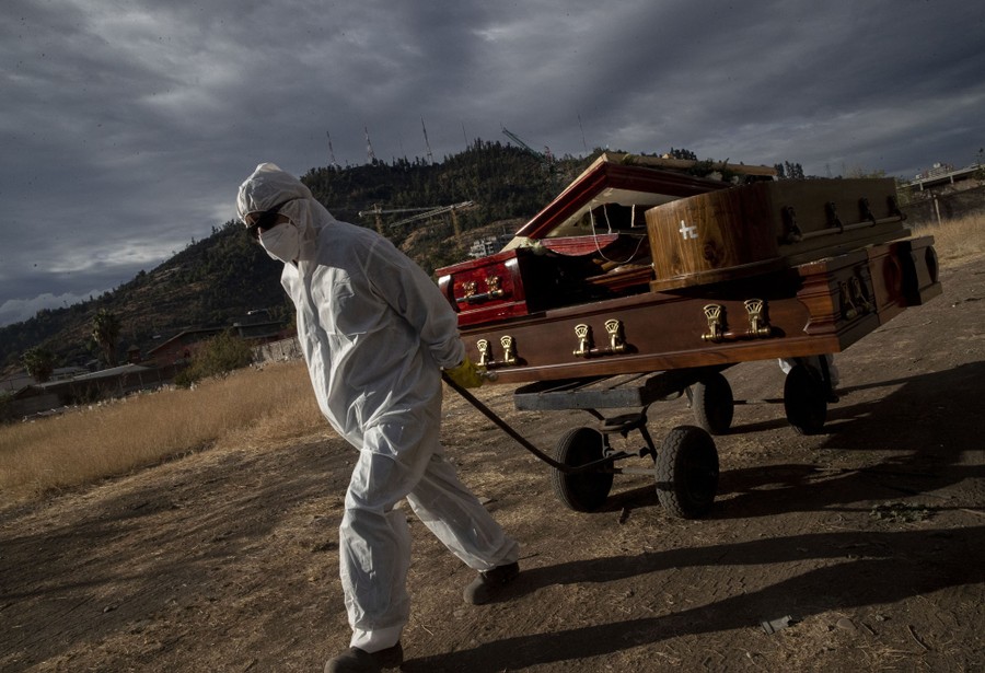 A funeral worker in protective gear pulls a cart carrying several empty coffins.