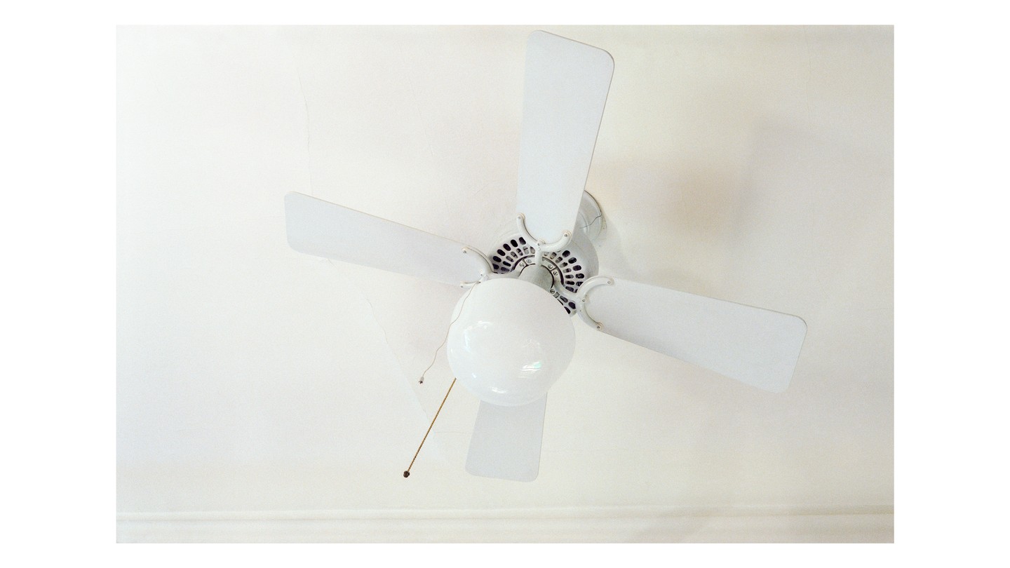 A gauzy photograph, closely cropped, of a white ceiling fan and an unlit bulb, on a white ceiling