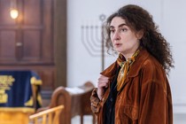 A woman with thick eyebrows and curly hair stands in front of an alter with a visible menorah.
