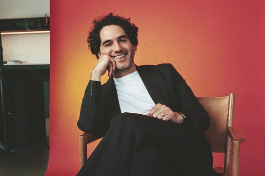 A color photograph of Benny Safdie seated in front of a red backdrop