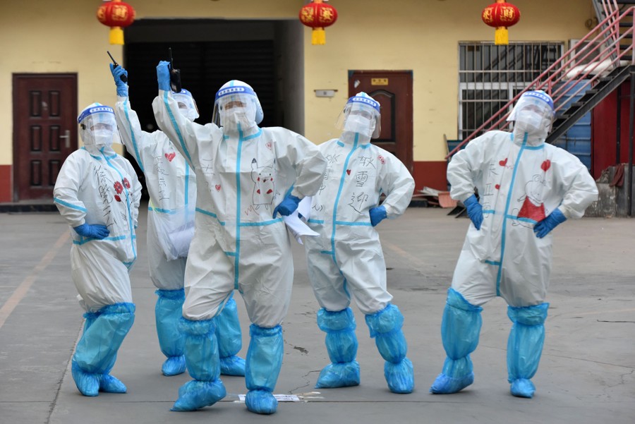 Medical workers in full protective gear dance and gesture in a parking lot.