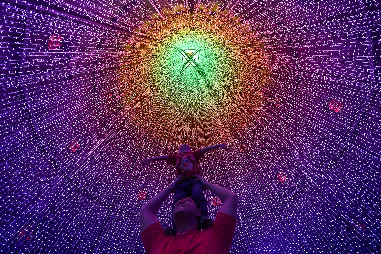 A man holds a small child high on his shoulders, posing beneath a tent of colored strings of Christmas lights.