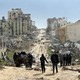 People walk on a dirt road past many damaged and destroyed buildings.