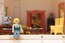 A Playmobil doll sits in her tiny, plastic living room. 