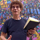 A curly haired artist reads from a book, in front of a mural he painted.