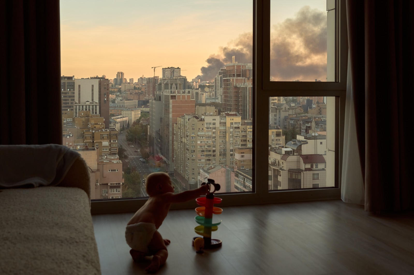 A toddler plays with a toy on an apartment floor, with a view through large windows of smoke rising from a fire in buildings in the distance, following a missile attack.