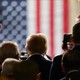 Donald Trump stands facing a U.S. flag that is blurred in the background.