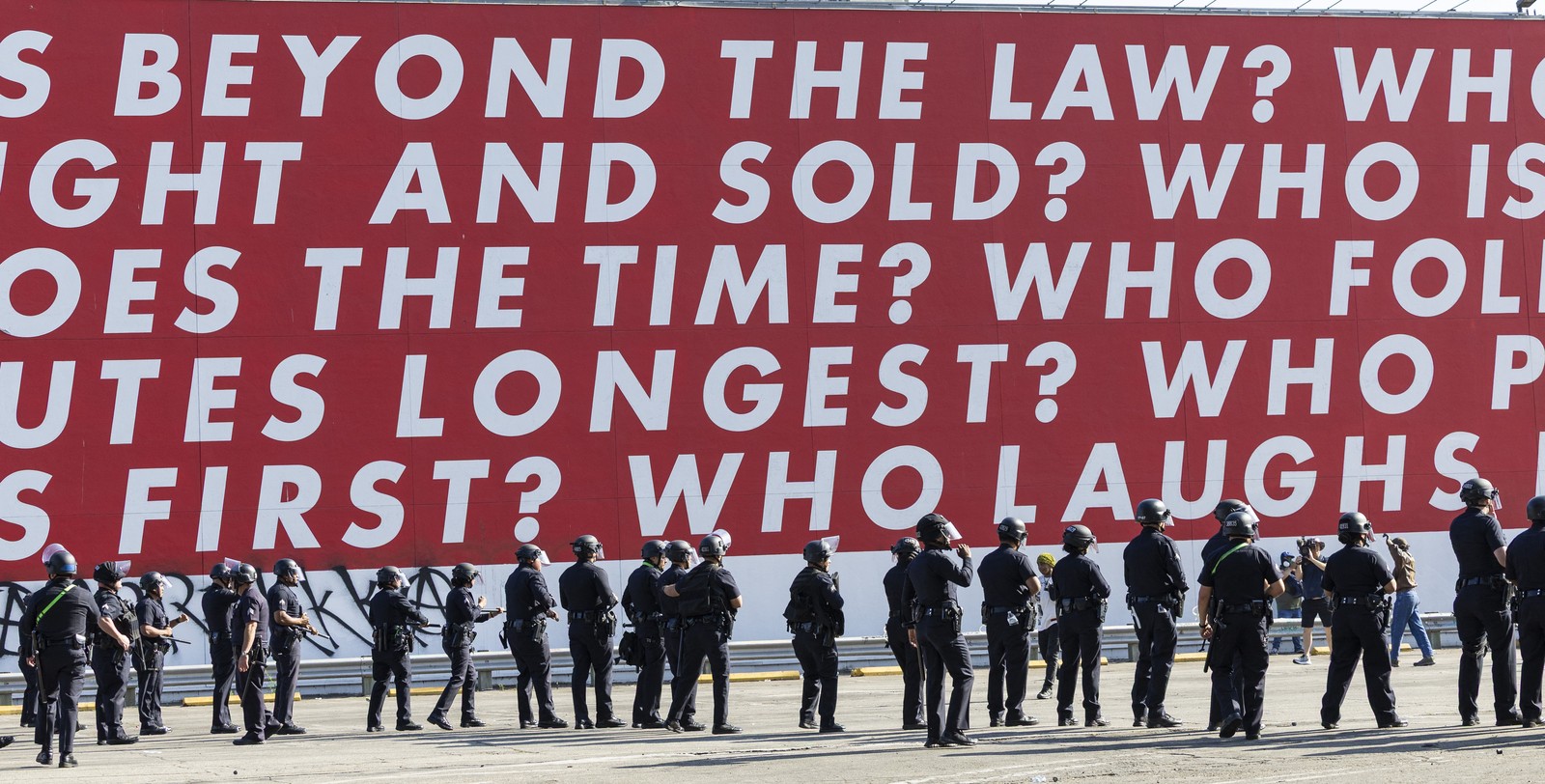 A line of riot police officers stand beneath a huge work of art on an outside wall, made up of text, reading in part 'beyond the law?'