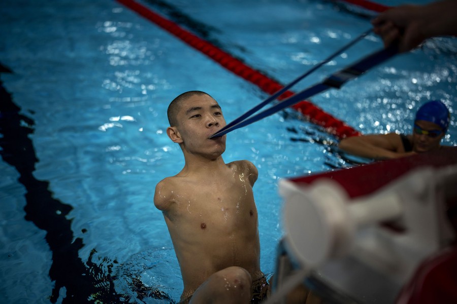 A swimmer with no arms holds a strap with his mouth, at the edge of a pool, readying to take off at the start of a practice race.