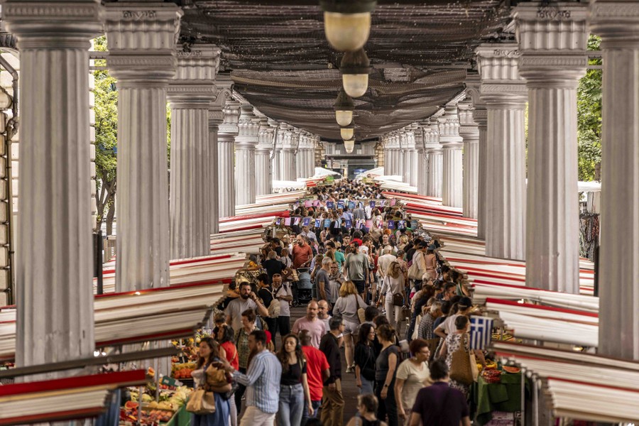 People shop in a crowded outdoor marketplace lined with ornate columns.