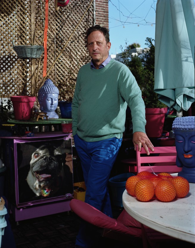 Binstock standing in a green sweater and blue pants with his hand on a pink chair outside.