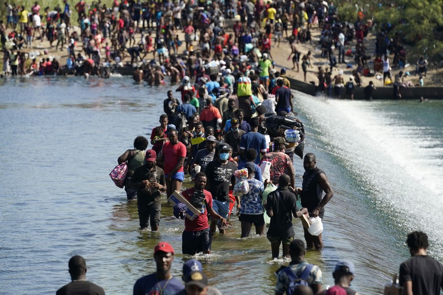 Hundreds of people line the banks of a river, many of them crossing by wading through shallow water along the top of a submerged weir dam.