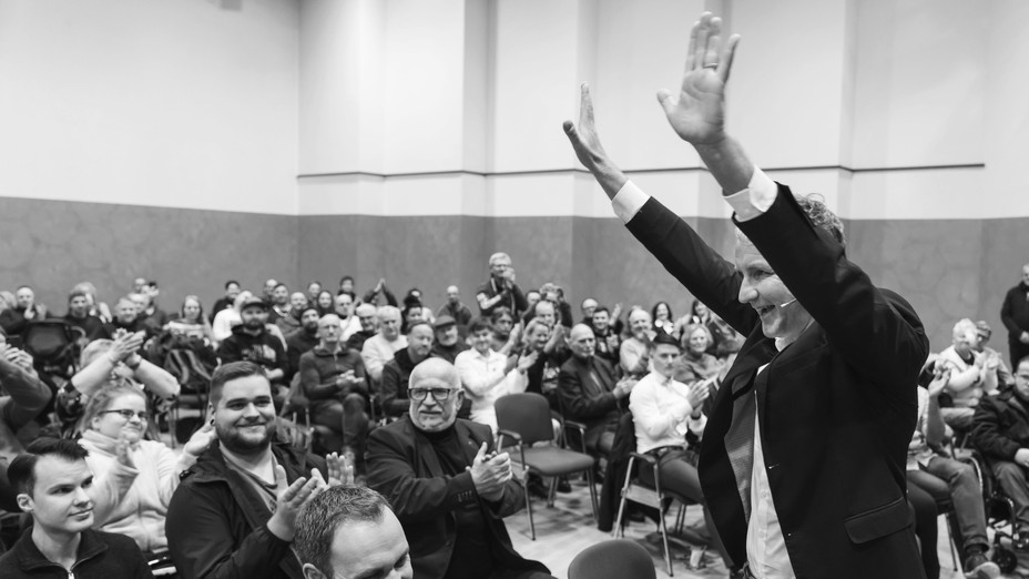 man stands raising his hands while facing a seated crowd