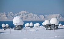Rows of white telecommunications domes stand on a snow-covered field before a distant view of mountains.