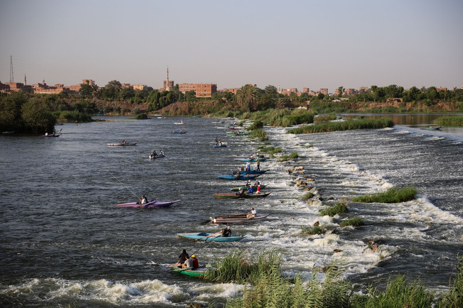 People in many small boats fish along a broad and shallow waterfall on a river.