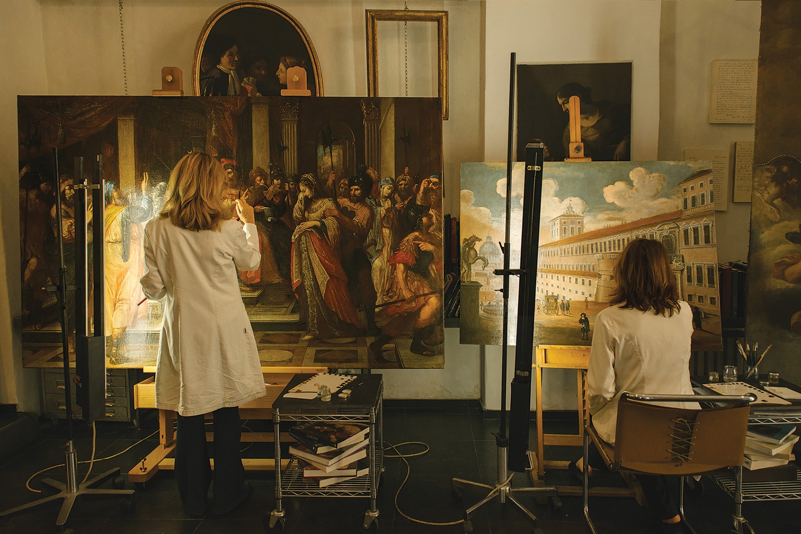 photo of two women from back, one sitting and one standing, while working to restore on large antique oil paintings on easels