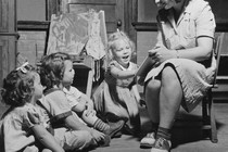 Black-and-white photo of a woman sitting in a chair reading a storybook to three kids seated on the floor