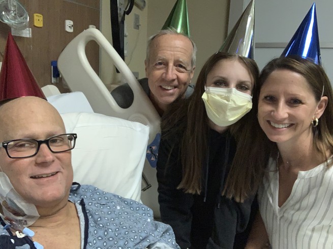 A man in a hospital bed smiles for the camera in a party hat. An older man, a young woman, and a middle-aged woman also wearing party hats smile from his bedside.