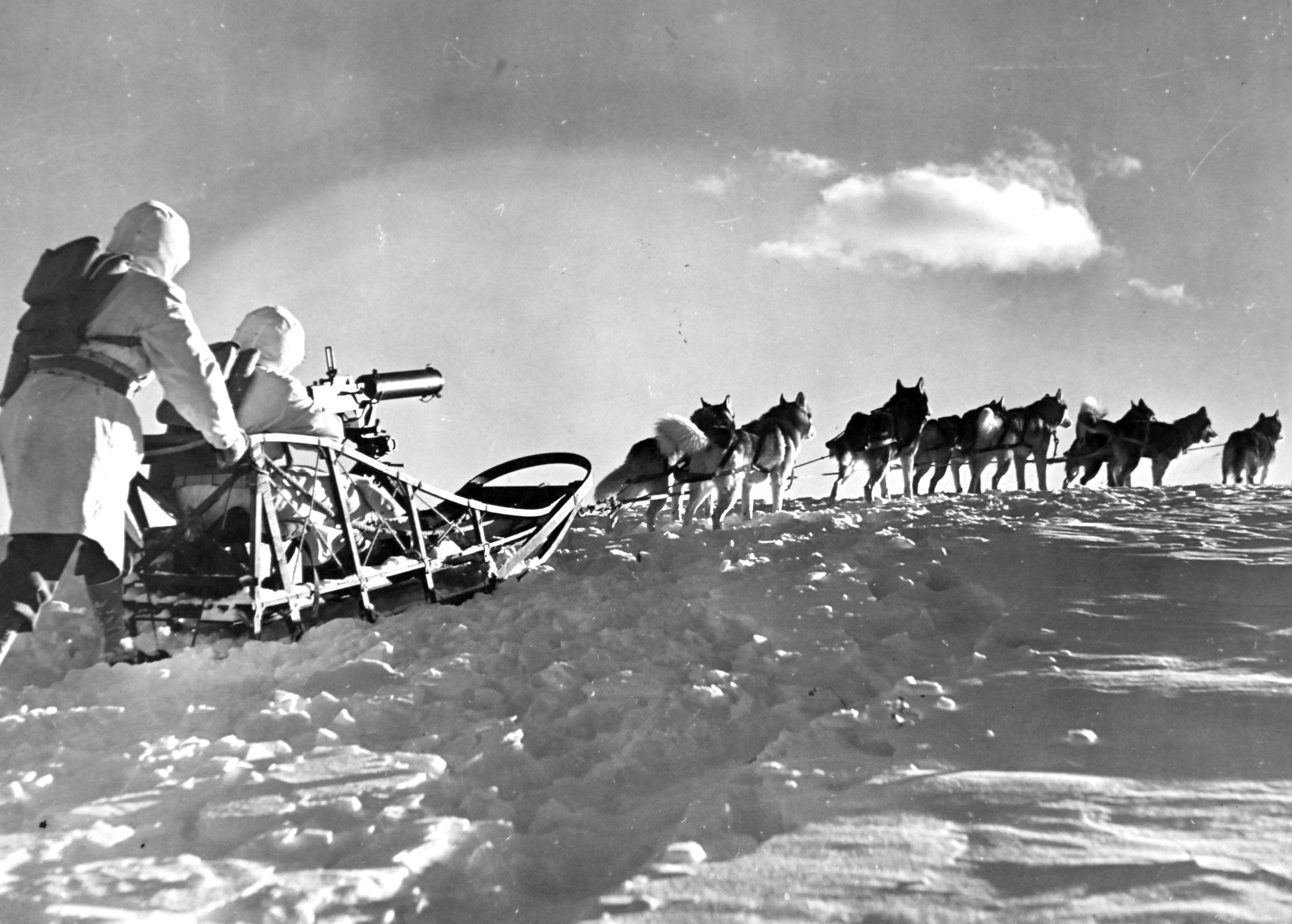Two soldiers wearing winter camouflage sit in and push a dog sled with a machine gun mounted on it, behind a team of sled dogs.