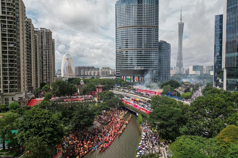 An elevated view of crowds of people lining a river in a city, where many dragon boats prepare for races