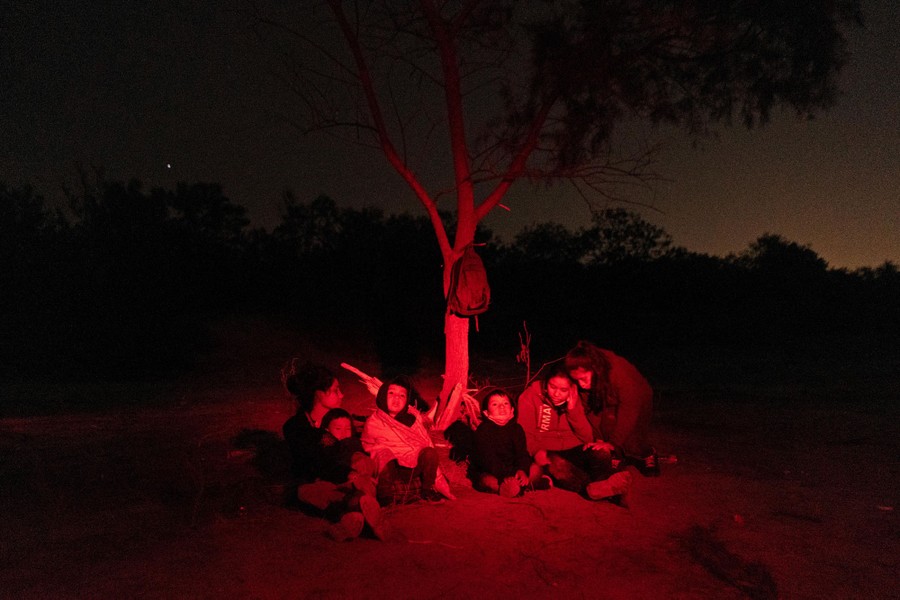 Six people, several of them children, sit on the ground beside a small tree at night, waiting.