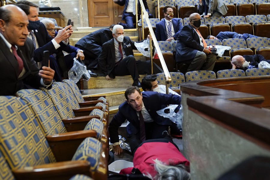 Several people lie on the floor, as others sit or crouch low, all looking apprehensive, in a seating gallery.