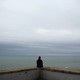 A person sitting on a low wall looking at the ocean under an overcast sky