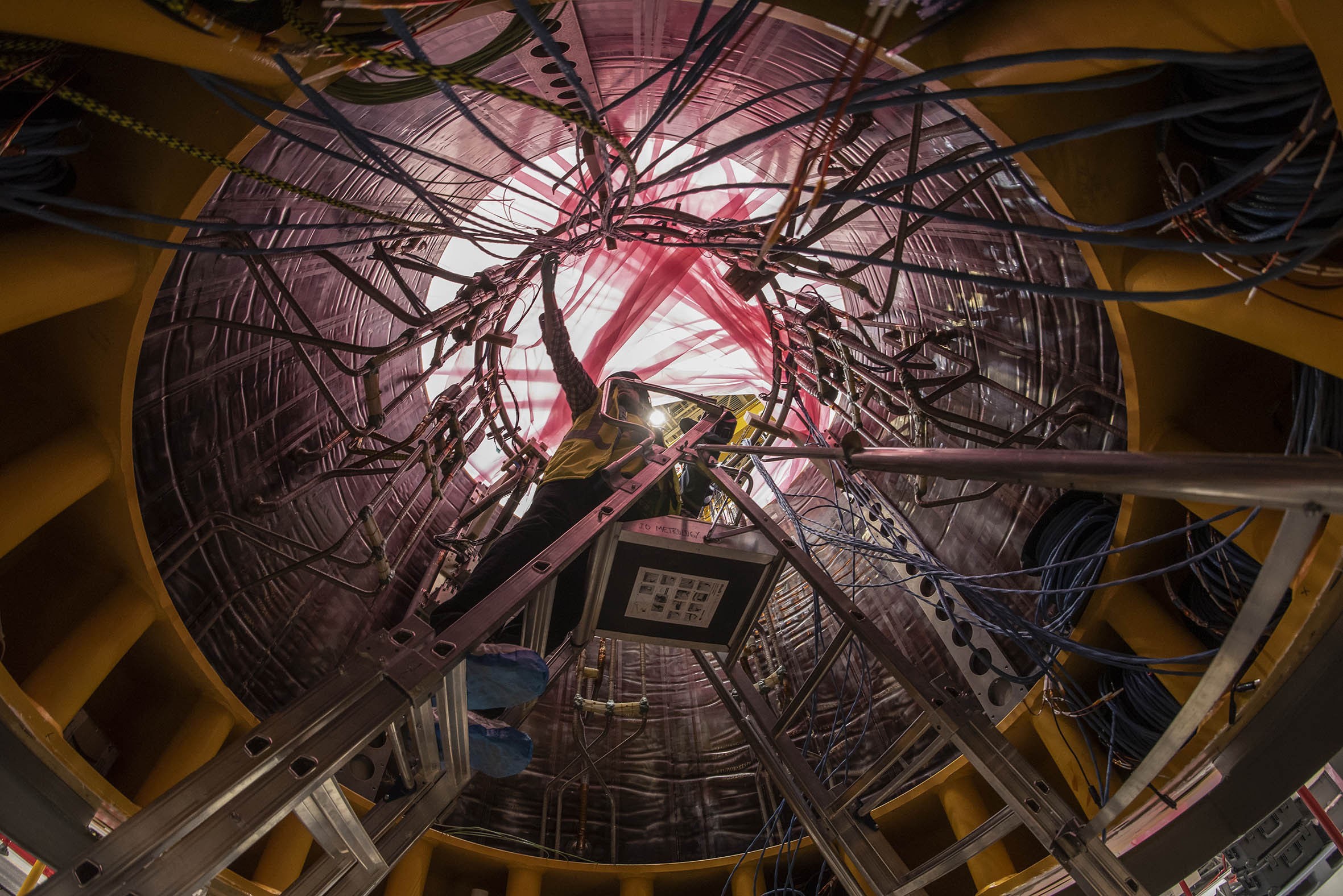 A view looking upward into the interior of a complicated cylindrical structure about 10 feet around; a worker on a ladder stands inside