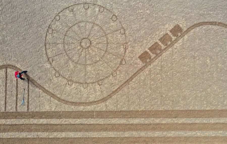 An artist creates a depiction of a rollercoaster and Ferris wheel in the sand of a beach.