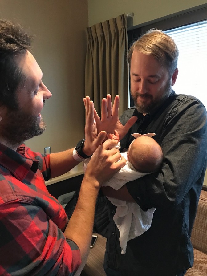 Andy and Gabe hold up a baby's hand to high five them in a hospital room