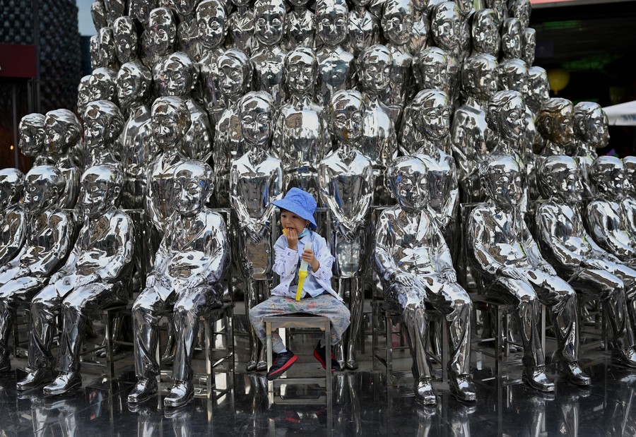 A boy sits amid dozens of silvery sculptures of seated men.