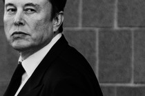 A black-and-white photo of Elon Musk in a suit and tie, looking back over his left shoulder.