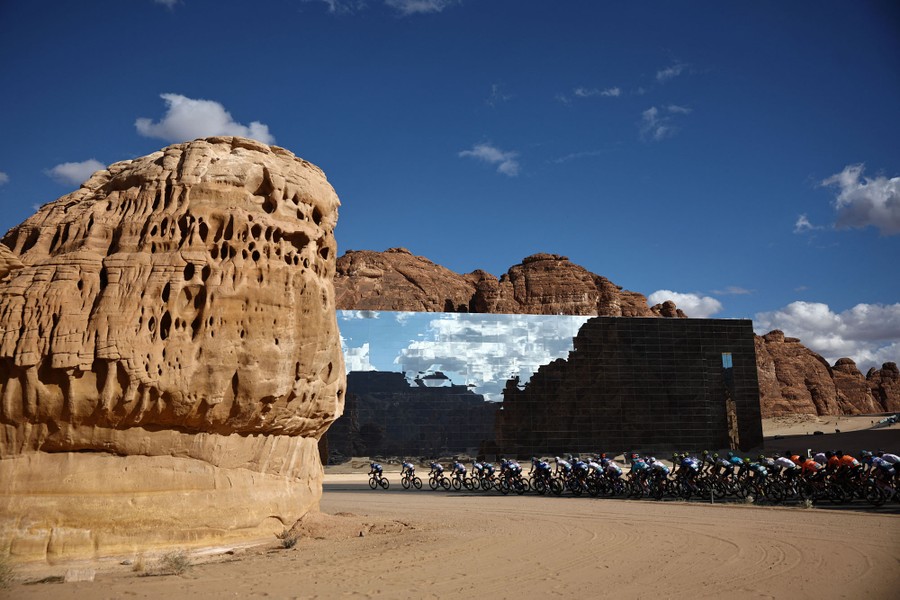 A pack of racing cyclists rides past a large mirrored building in a desert.