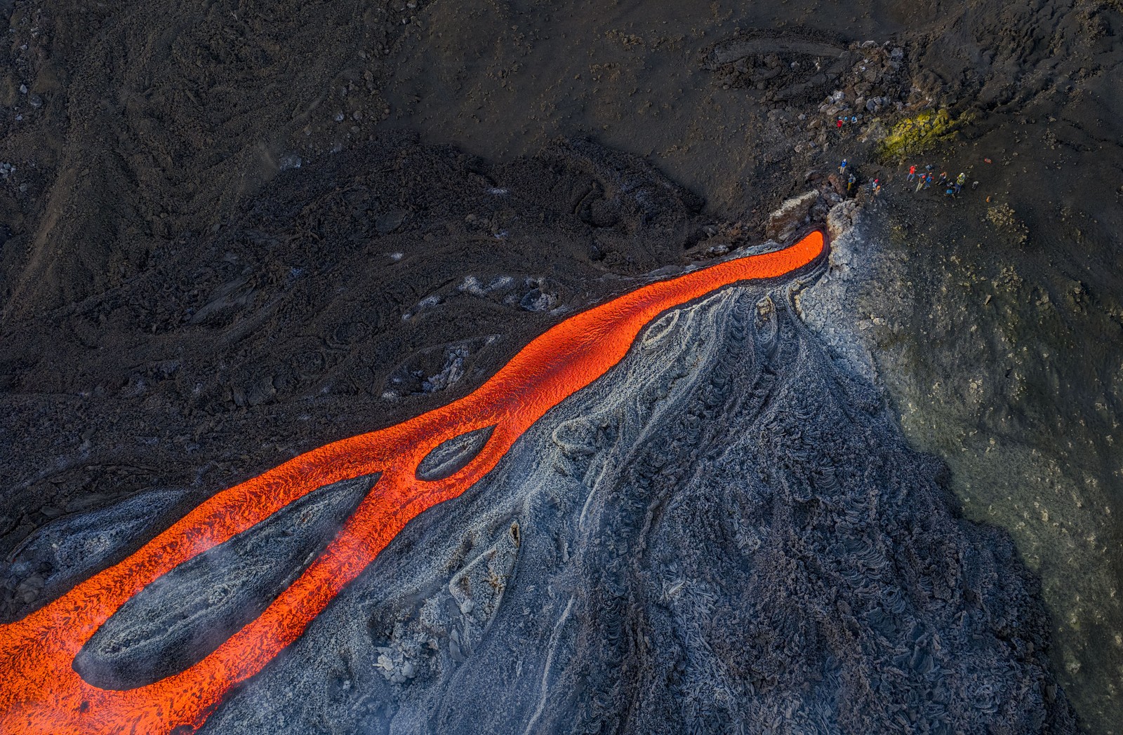 An aerial view of people standing near the outlet of a lava flow on the side of a volcano