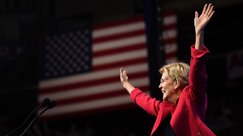 Elizabeth Warren waves to a crowd in front of an American flag.