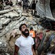 A man reacts, standing beside a huge pile of rubble following an air strike. Workers and onlookers stand nearby and atop the rubble.