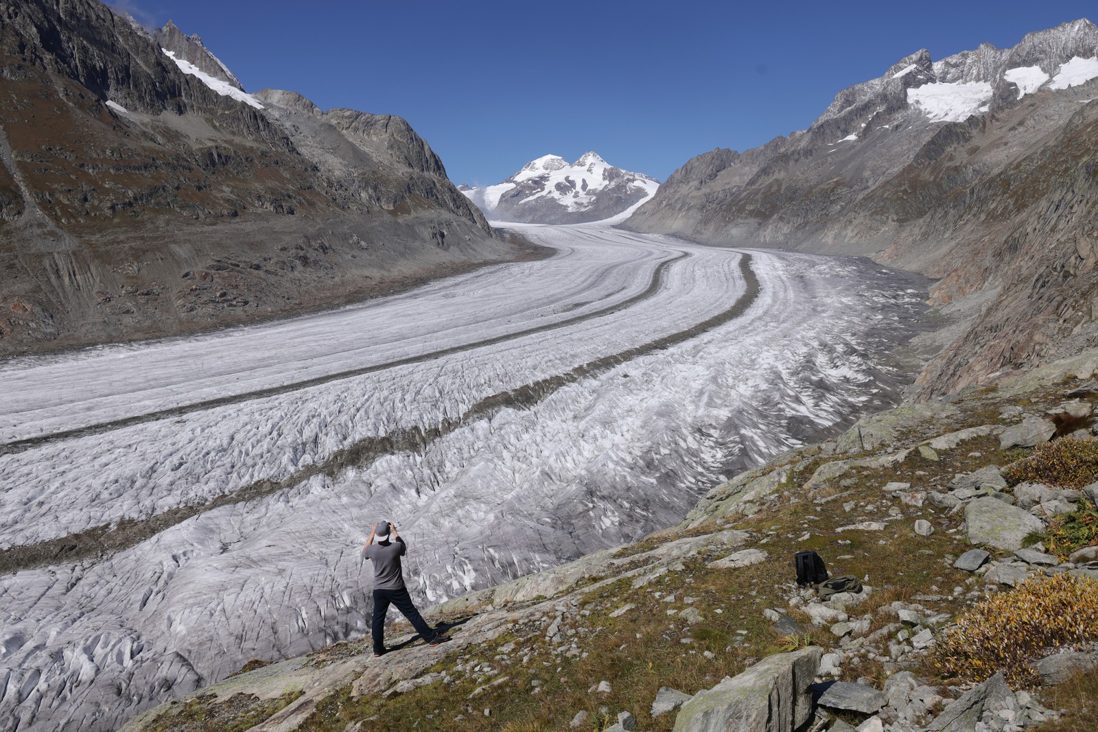 A hiker stands on a rocky slope, looking out over a broad glacier.