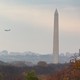 A plane flies in the sky over trees with multicolored leaves as the Washington Monument stands in the background.