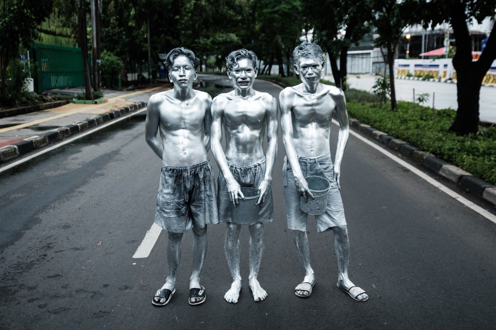 Three street performers, completely covered in silver paint, stand side-by-side on a road, posing.