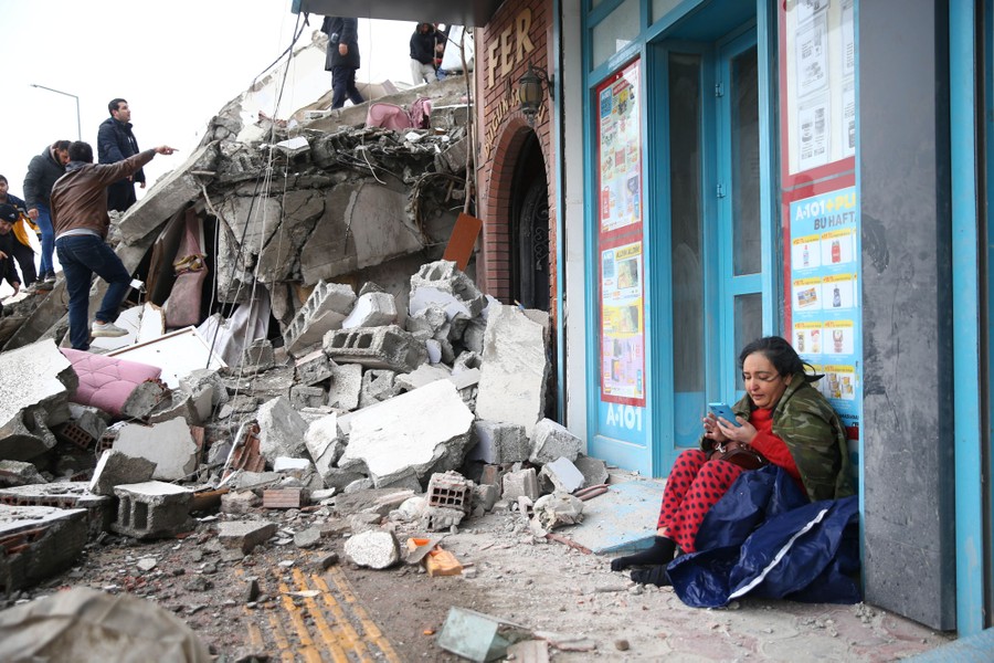 A person sits on a sidewalk, using their phone, as search-and-rescue operations are carried out at a collapsed building nearby.