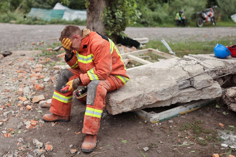 A worker in an orange uniform sits on a piece of rubble, holding a hand to their head.