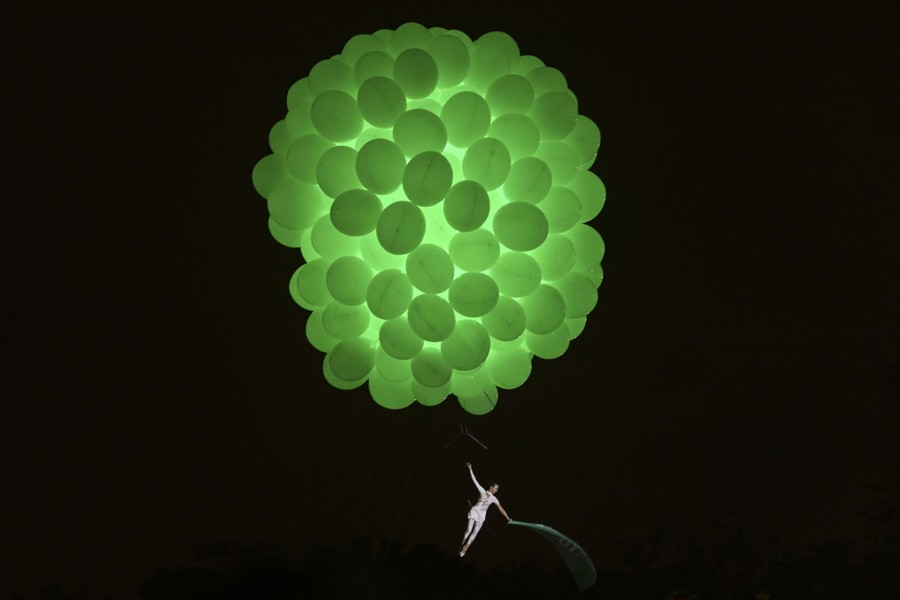 A performer hangs beneath a huge cluster of glowing green balloons.