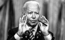 President Joe Biden speaks into a microphone with his hands held up to frame the lower half of his face.