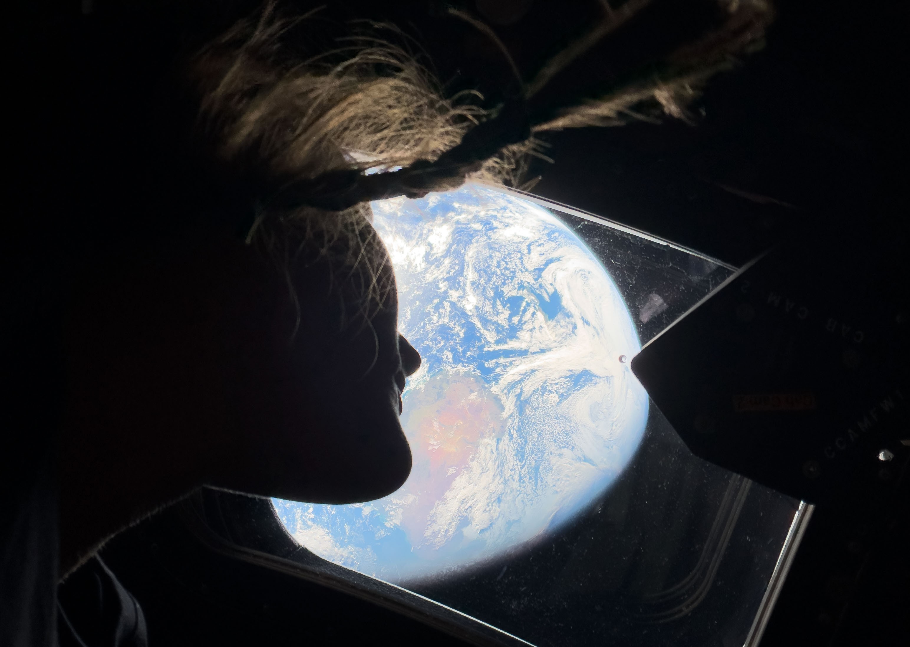 An astronaut, seen in profile, with the brightly-lit Earth in the background, seen through a window